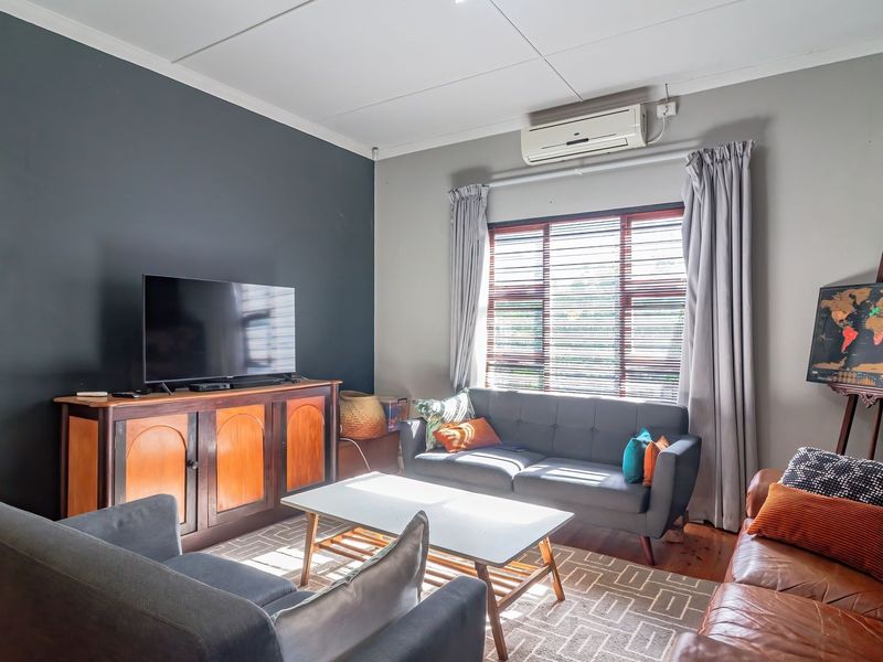 A comfortable living room featuring timber flooring, an air conditioning unit, and large windows with venetian blinds, creating a bright and functional space.
