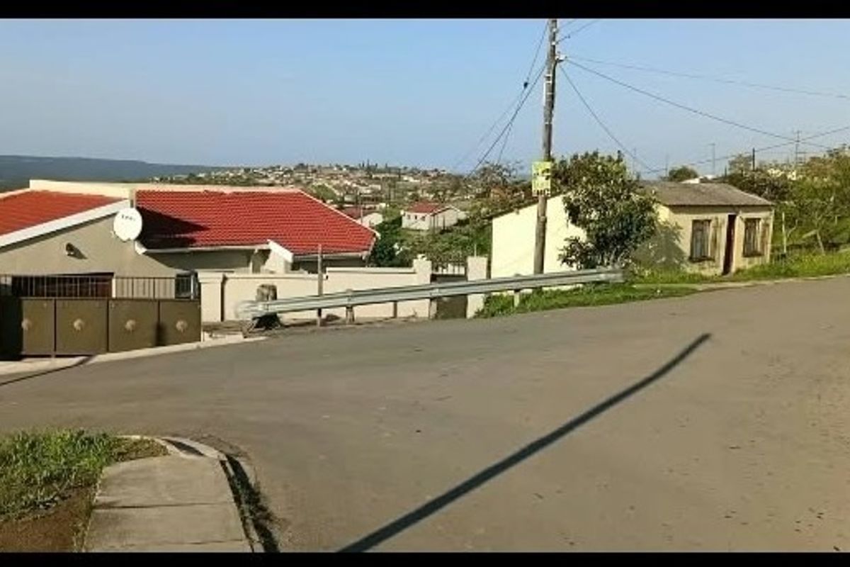 A street-level view of the property showing the asphalt road, perimeter wall, metal gate, and surrounding residential neighborhood.