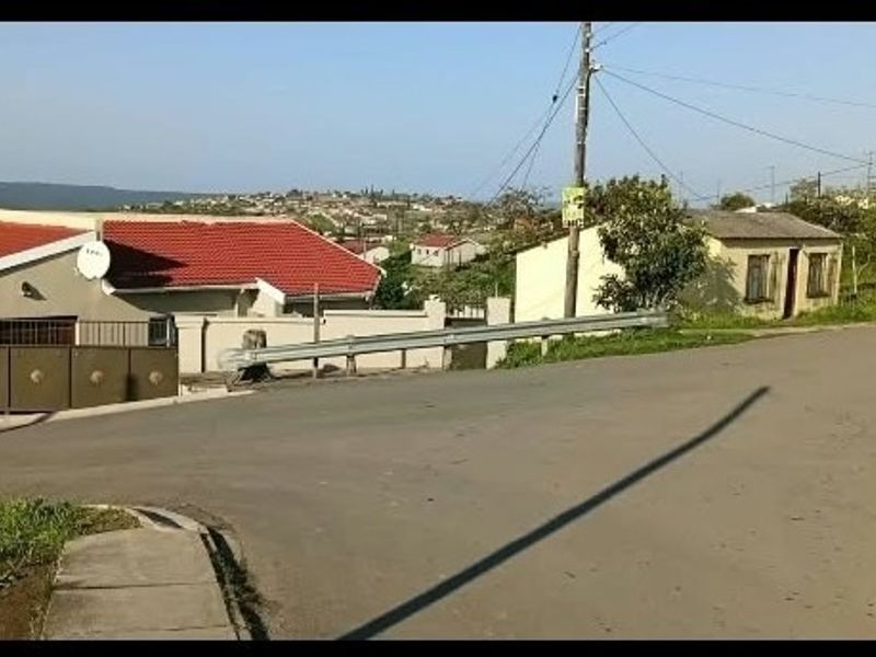 A street-level view of the property showing the asphalt road, perimeter wall, metal gate, and surrounding residential neighborhood.