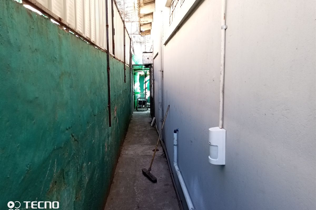 A narrow exterior side passage or rear service area featuring a concrete walkway, boundary wall, and external utility conduits.