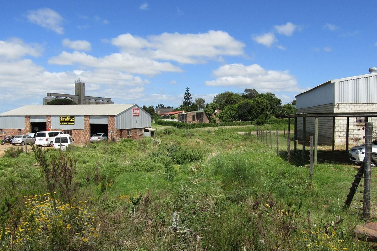 A view of a vacant residential plot featuring overgrown grass and surrounding industrial and residential structures.