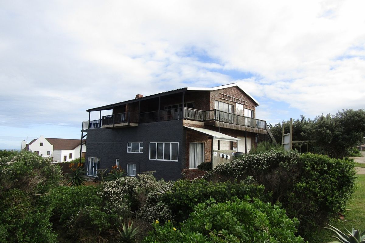 A multi-level brick house featuring a large upper balcony, sliding glass doors, and a corrugated roof, set within a landscaped garden area.