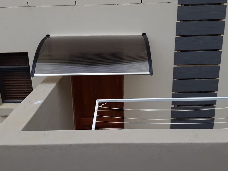 Rear exterior view of the flat featuring a timber entrance door, a protective polycarbonate awning, and a fixed wall-mounted washing line.