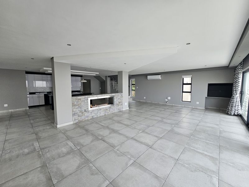 Spacious open-plan living area featuring large format floor tiles, a central stone-clad fireplace, and a modern kitchen with sleek cabinetry.