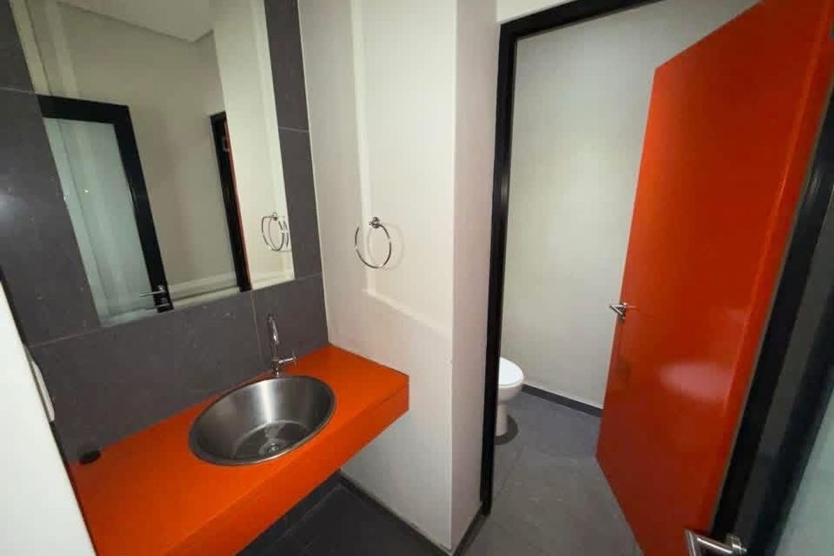 Modern office restroom featuring a vibrant orange vanity with a stainless steel basin, dark tiled flooring, and a separate toilet cubicle.
