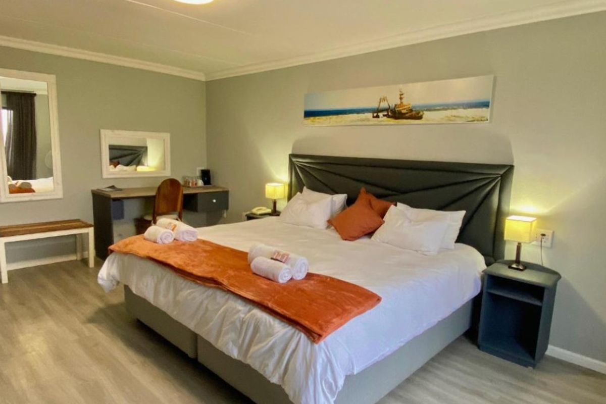 A well-maintained hotel bedroom featuring light wood-look flooring, neutral wall tones, and a built-in desk area with a wall-mounted mirror.