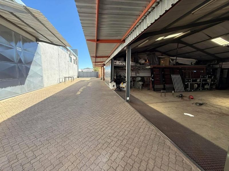 Industrial exterior area featuring a paved driveway, covered loading bay with steel structure, and access to a large warehouse space.