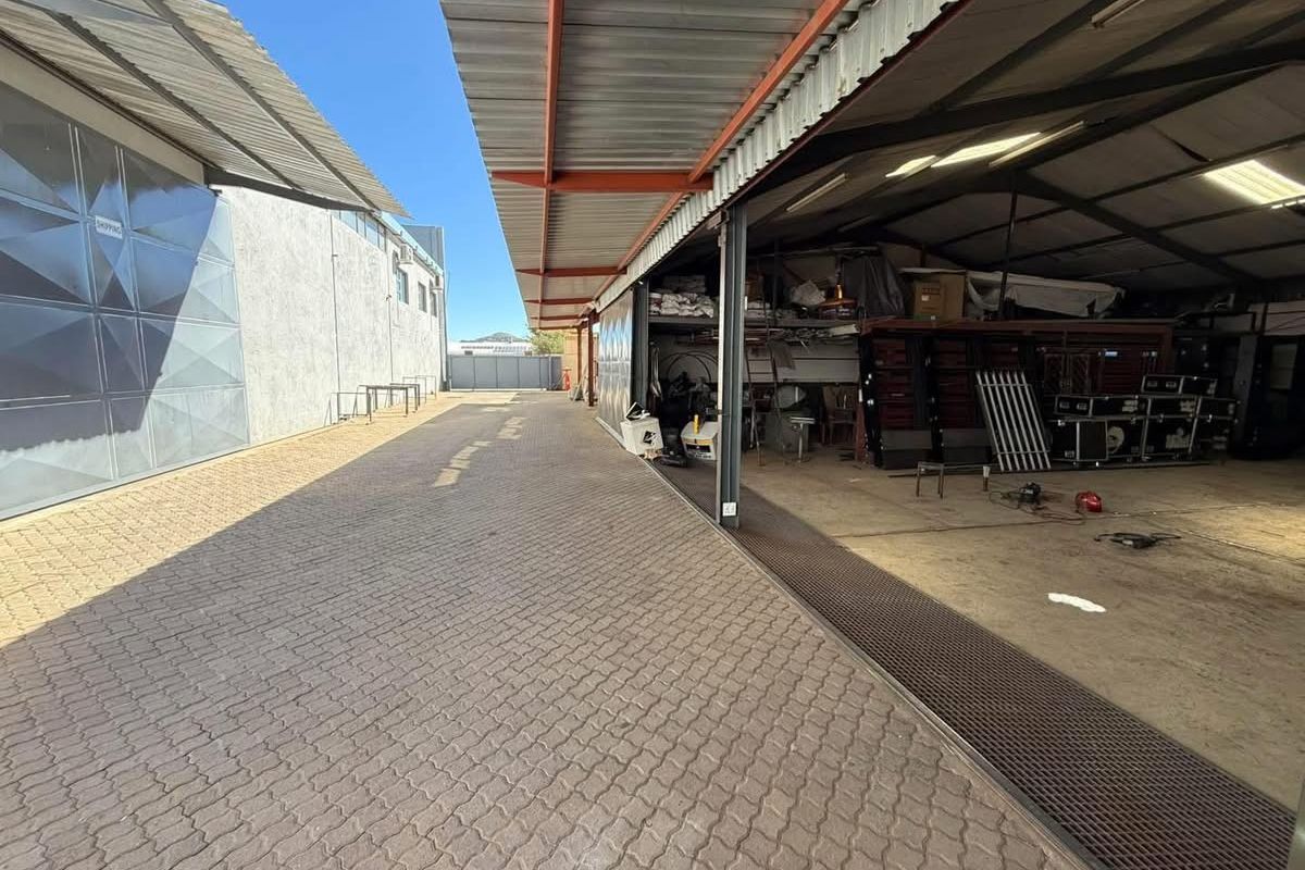 Industrial exterior area featuring a paved driveway, covered loading bay with steel structure, and access to a large warehouse space.