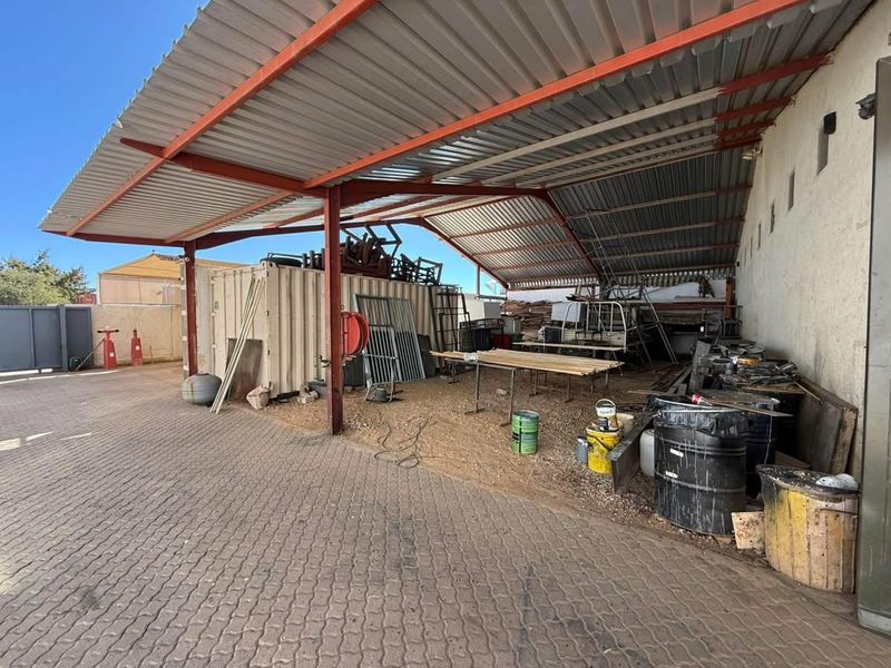 Covered industrial utility area featuring a corrugated metal roof, steel support structure, and paved ground, suitable for storage or workshop activities.