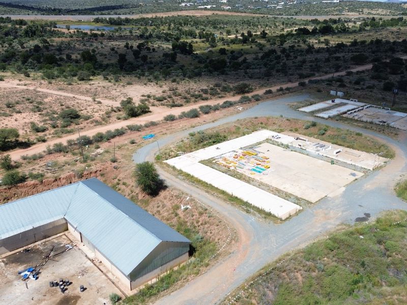 Aerial view of an industrial site featuring a large metal-roofed structure, expansive concrete slabs, and surrounding undeveloped land with gravel access roads.