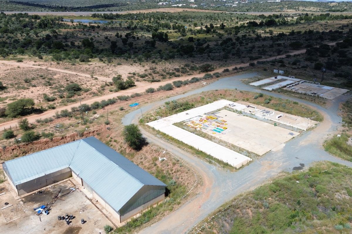 Aerial view of an industrial site featuring a large metal-roofed structure, expansive concrete slabs, and surrounding undeveloped land with gravel access roads.