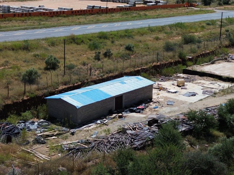 Aerial view of an industrial site featuring a small structure with a blue metal roof, concrete slab, and surrounding open yard space.