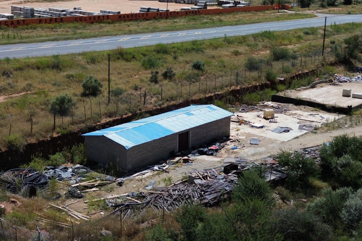 Aerial view of an industrial site featuring a small structure with a blue metal roof, concrete slab, and surrounding open yard space.