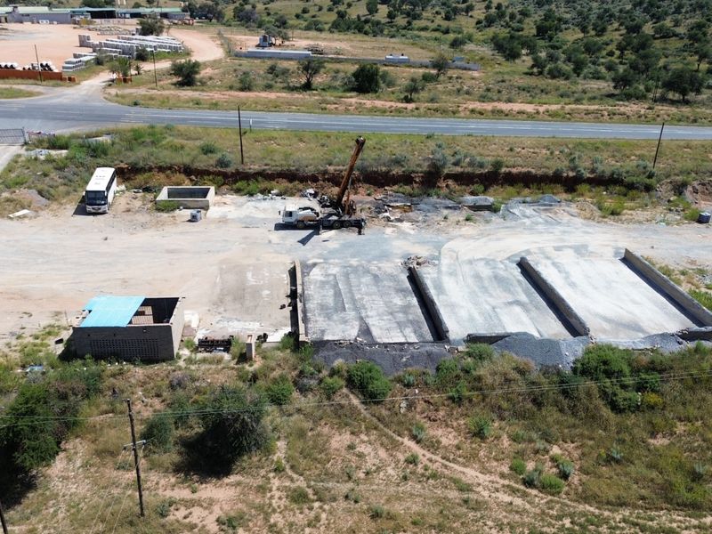 Aerial view of an industrial site featuring concrete loading bays, a small utility structure, and a large gravel yard area adjacent to a main road.