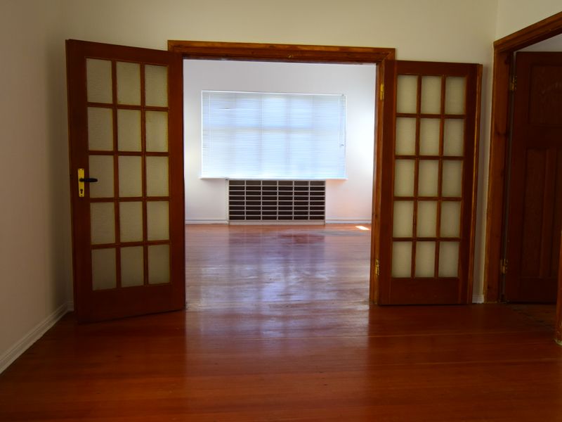 An empty living area featuring polished timber flooring, classic timber-framed french doors, and a built-in shelving unit under a large window.