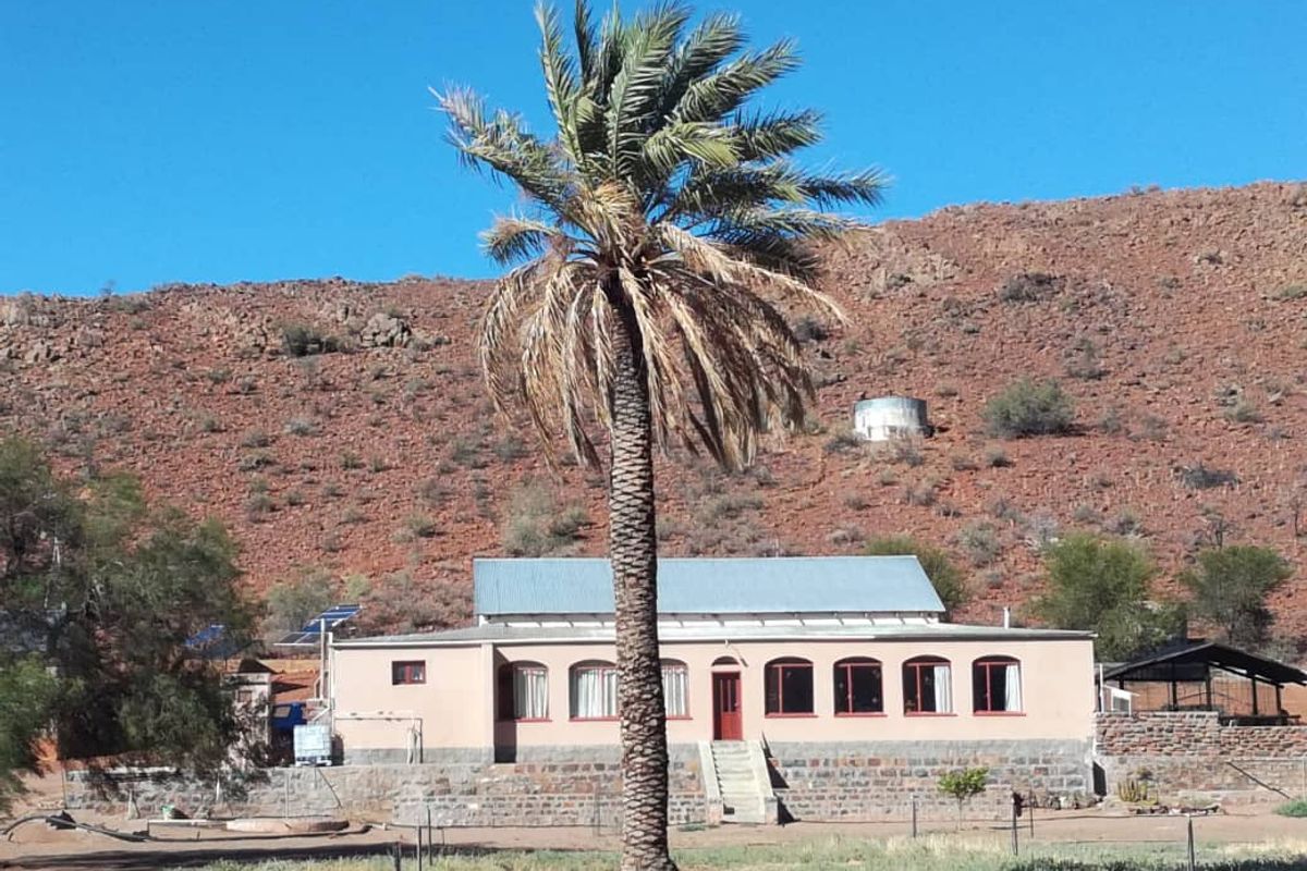 A farmstead exterior featuring a single-story dwelling with a metal roof, stone base, and solar panels, set against a rugged mountain landscape.