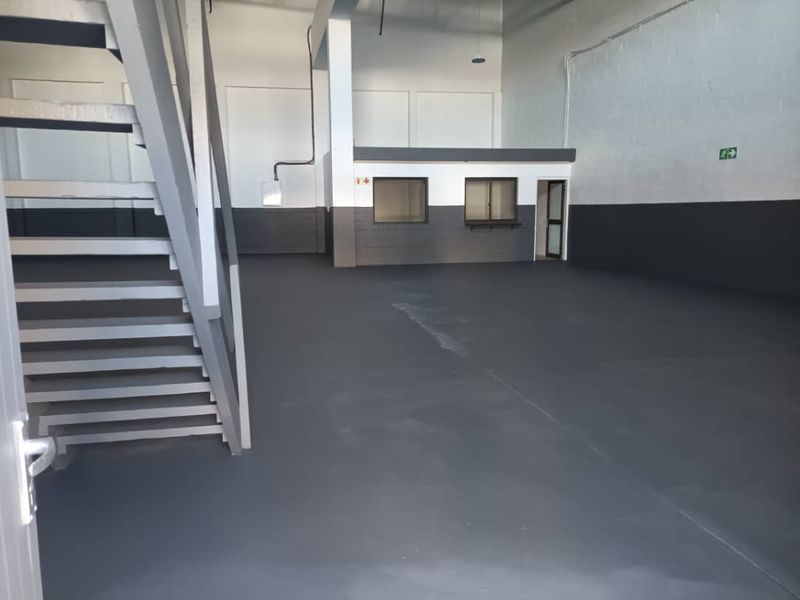 Spacious industrial warehouse unit featuring a painted concrete floor, internal office partition with service windows, and a metal staircase leading to an upper level.