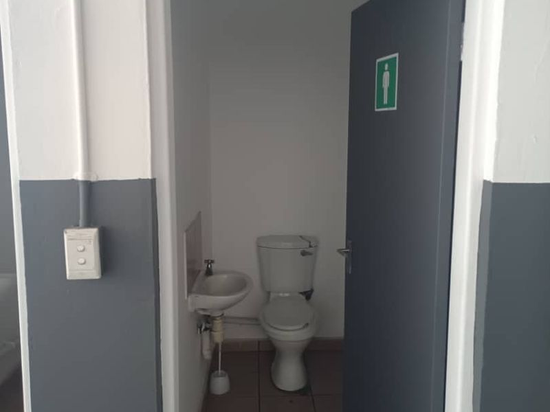 A functional industrial-style restroom featuring a toilet, wall-mounted basin, and tiled flooring, accessed via a single door.