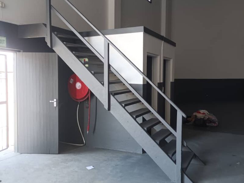 Industrial warehouse interior featuring a metal staircase leading to a mezzanine level, concrete flooring, and a wall-mounted fire hose reel.