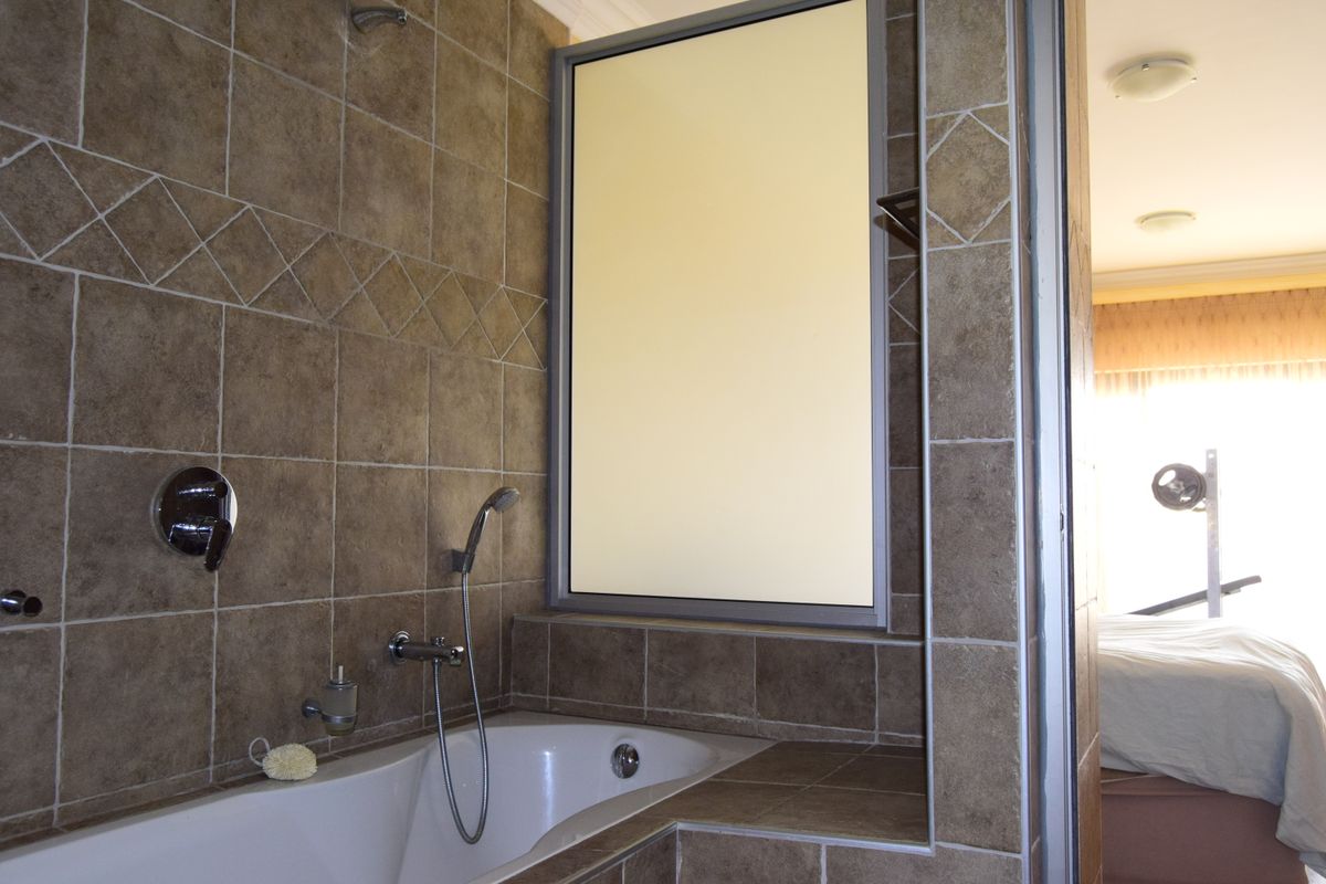 A bathroom featuring a built-in bathtub with a handheld shower fixture, tiled walls, and a frosted glass window, adjacent to a bedroom area.