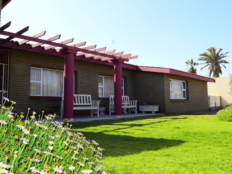 A covered patio area featuring a pergola structure, brick exterior walls, and paved flooring, overlooking a well-maintained garden.
