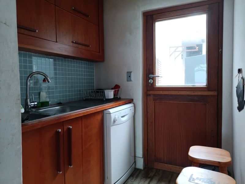 A functional scullery area featuring wooden cabinetry, a stainless steel sink, tiled backsplash, and direct access to the exterior via a wooden door.