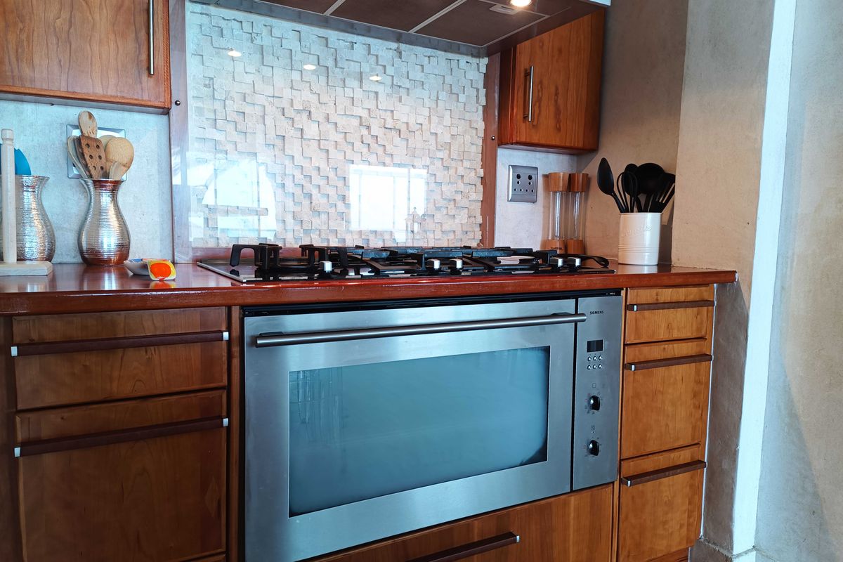 A well-maintained kitchen featuring wooden cabinetry, a built-in oven, gas cooktop, extractor fan, and a unique textured stone splashback.