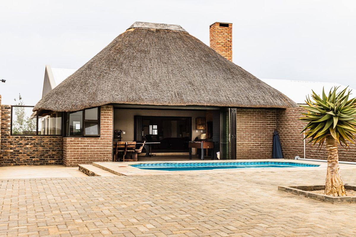 Exterior view of a thatched-roof dwelling featuring a swimming pool, paved patio area, and brick facade with large folding doors.