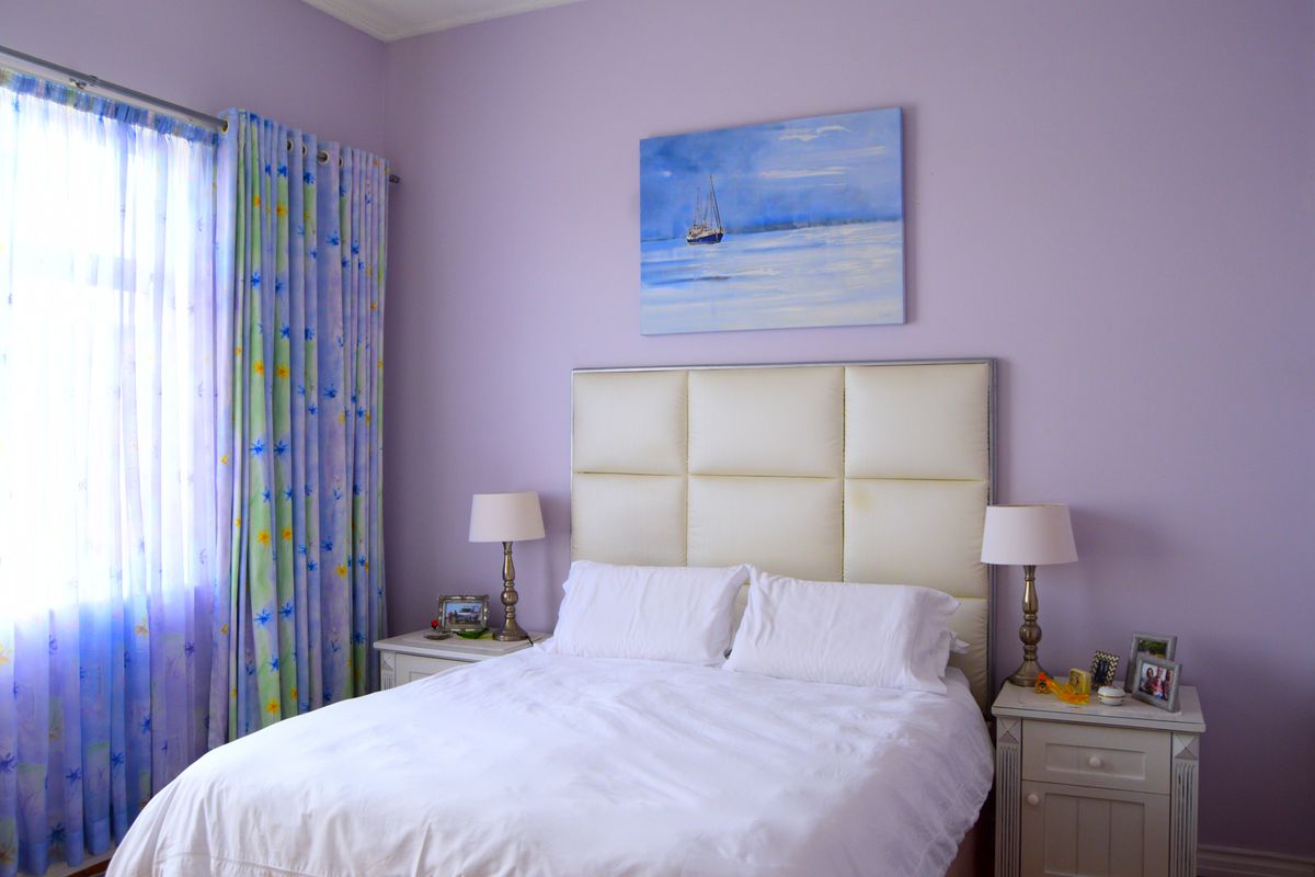 A bedroom featuring lavender-painted walls, a large window with patterned curtains, and a neutral-toned upholstered headboard.