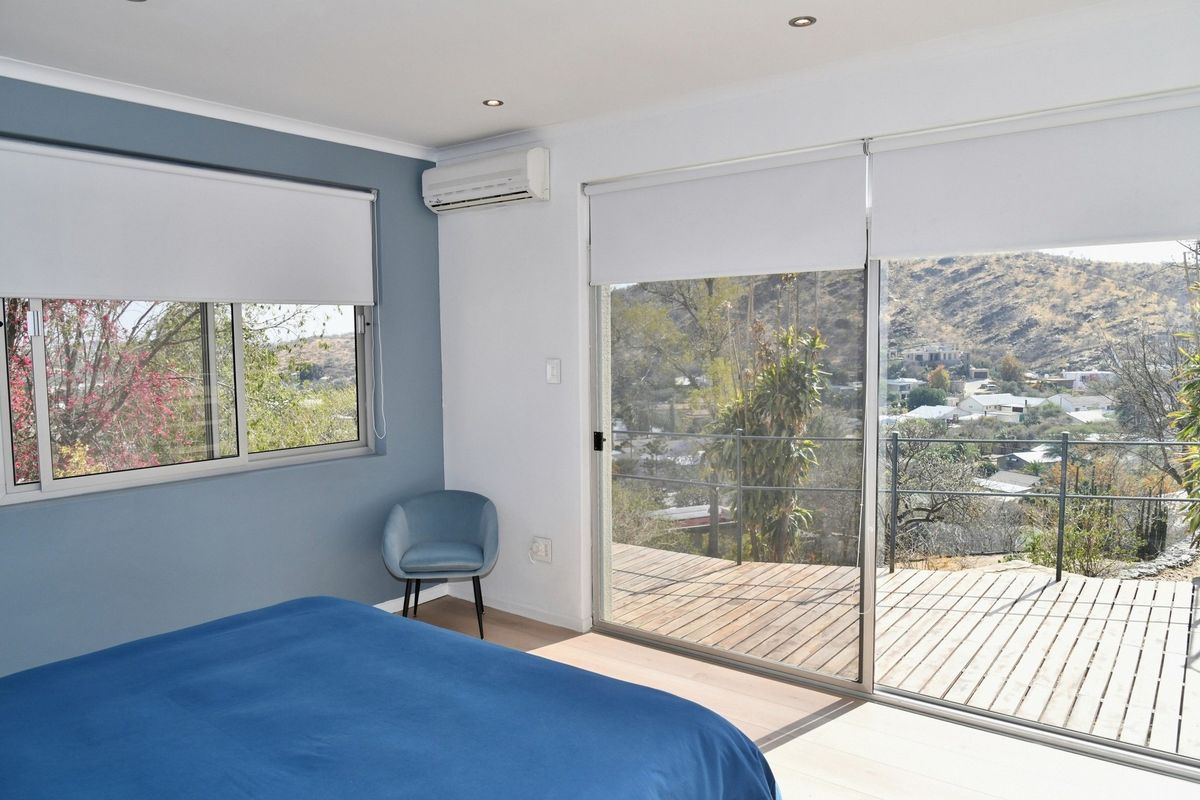 Bright bedroom featuring large sliding glass doors opening onto a wooden deck with scenic hillside views, complemented by a wall-mounted air conditioning unit.