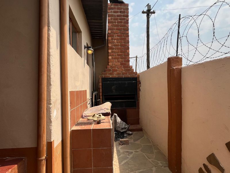 Outdoor patio area featuring a built-in brick braai, tiled prep counter, and stone paving, enclosed by a perimeter wall with security fencing.