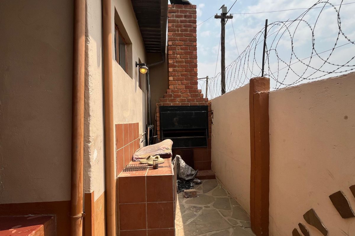 Outdoor patio area featuring a built-in brick braai, tiled prep counter, and stone paving, enclosed by a perimeter wall with security fencing.