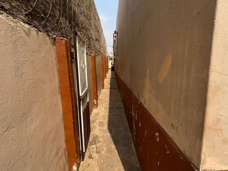 A narrow side passage featuring stone paving, boundary walls with security fencing, and a metal security gate.