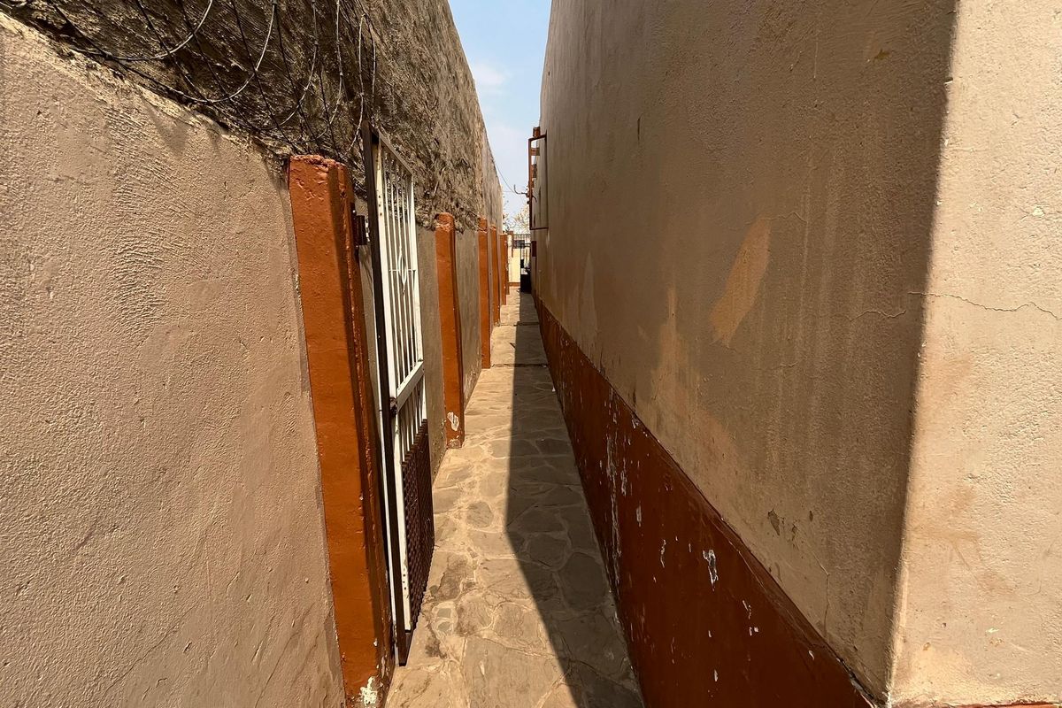 A narrow side passage featuring stone paving, boundary walls with security fencing, and a metal security gate.