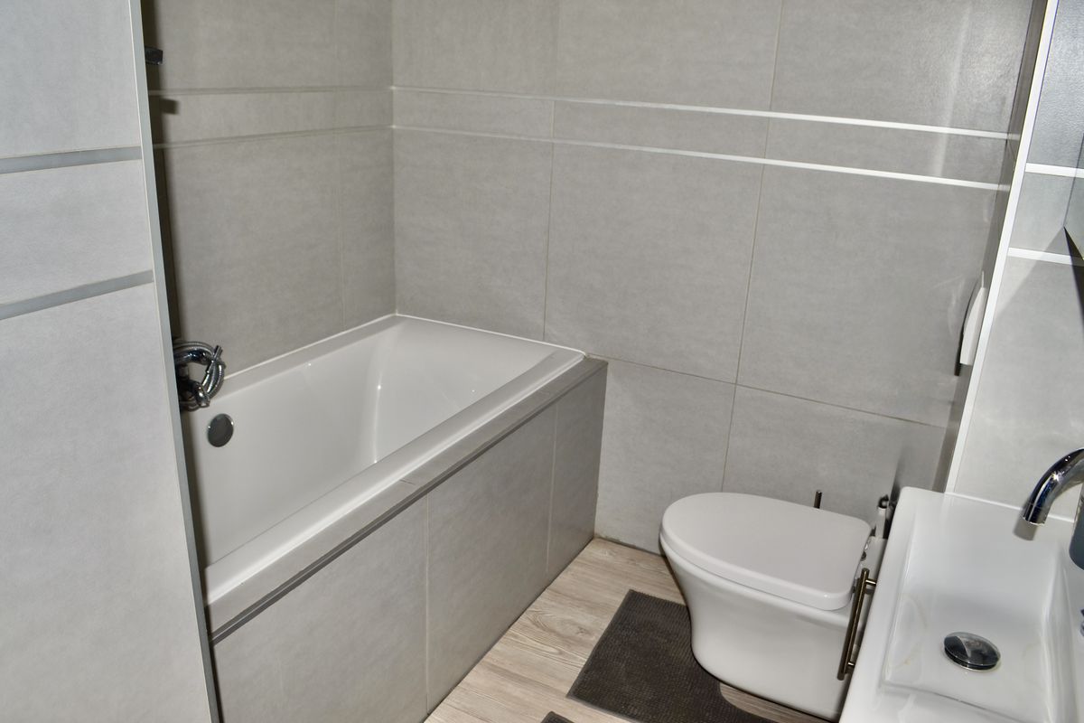 Modern bathroom featuring a built-in bathtub, wall-mounted toilet, and a sleek vanity basin with contemporary grey wall tiling and light wood-look flooring.