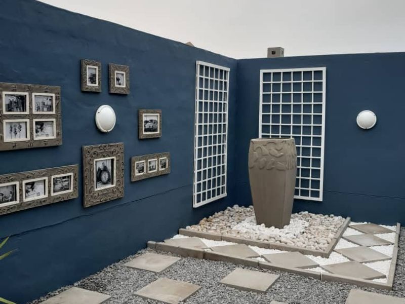 A landscaped garden area featuring a blue boundary wall, decorative trellises, a stone water feature, and a gravel path with concrete pavers.
