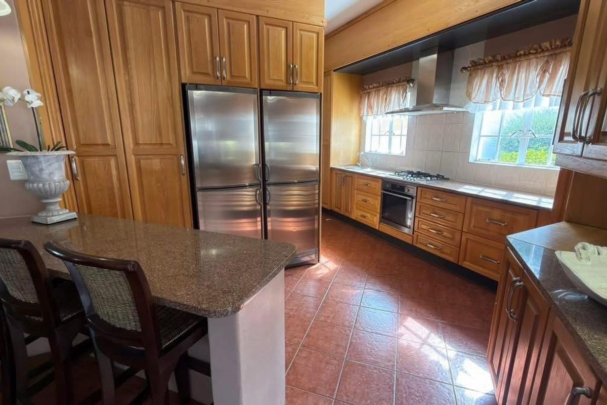 A spacious kitchen featuring warm-toned wooden cabinetry, granite countertops, stainless steel appliances, and tiled flooring with a functional breakfast bar area.