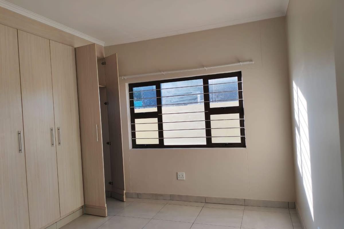 A bright bedroom featuring built-in wooden wardrobes, tiled flooring, and a window with security bars.
