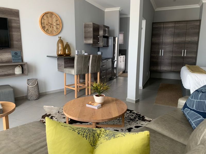 An open-plan apartment living area featuring a compact kitchenette with built-in cabinetry, tiled flooring, and a neutral color palette.