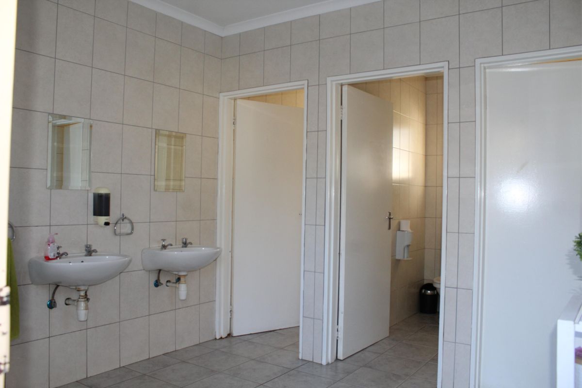 A functional bathroom area featuring dual wall-mounted basins, tiled walls and flooring, and multiple doorways leading to private toilet cubicles.