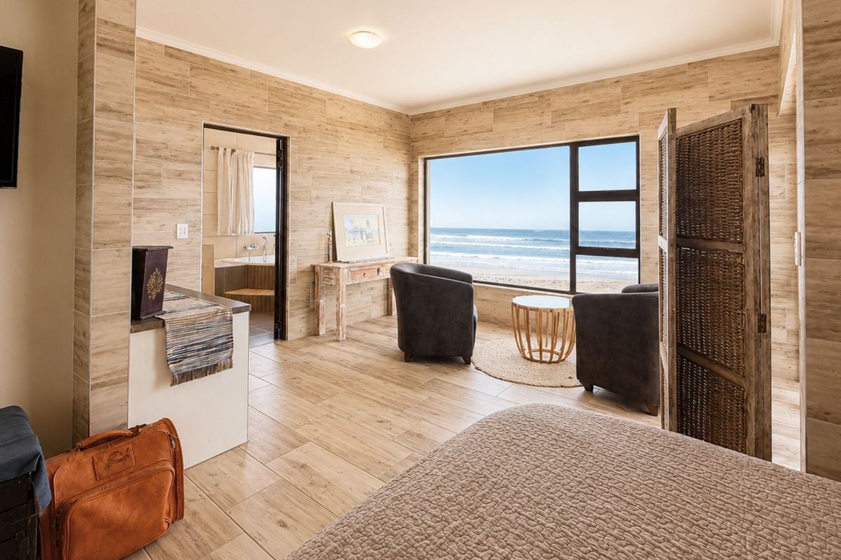A bright bedroom featuring wood-look tile flooring and wall cladding, large windows with direct ocean views, and an en-suite bathroom.
