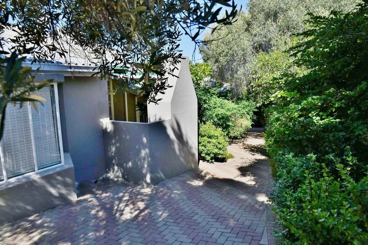 A paved pathway leads alongside the exterior of the house, surrounded by established garden greenery and mature trees.