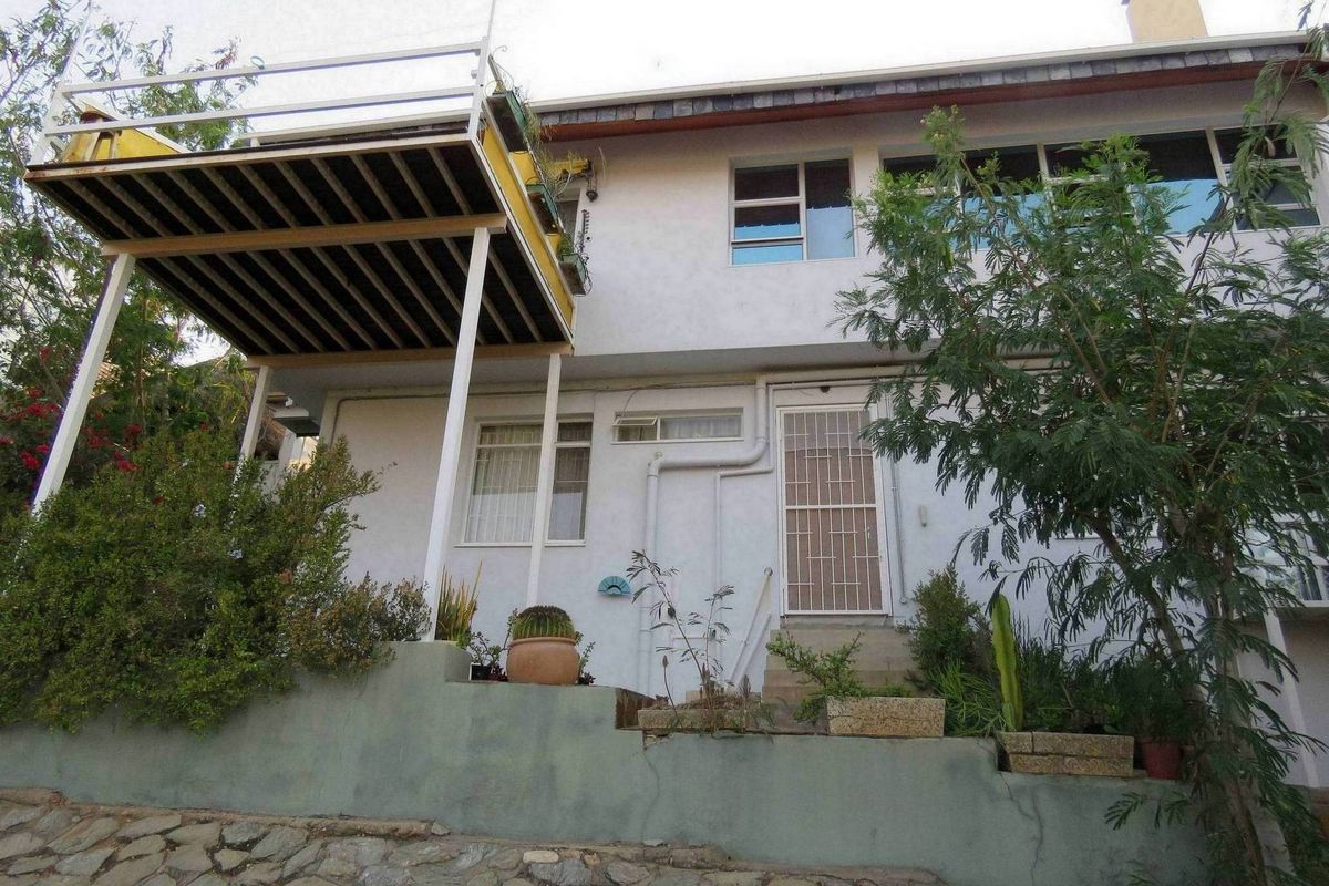 Rear exterior view of the house featuring a raised balcony, stone retaining wall, and established garden area.