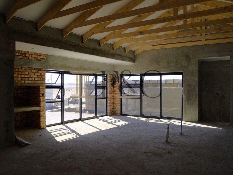 An unfinished open-plan living area featuring exposed timber roof trusses, a brick fireplace, and large aluminium windows with views of the exterior.