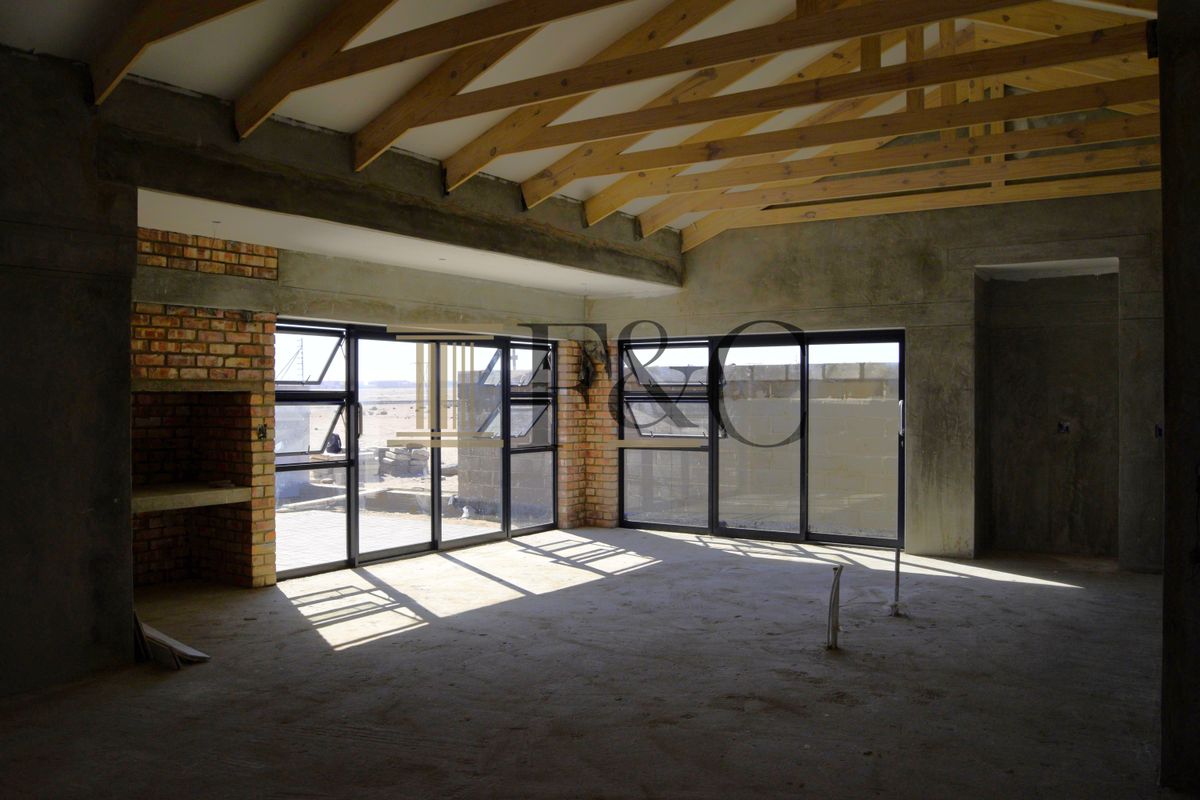 An unfinished open-plan living area featuring exposed timber roof trusses, a brick fireplace, and large aluminium windows with views of the exterior.