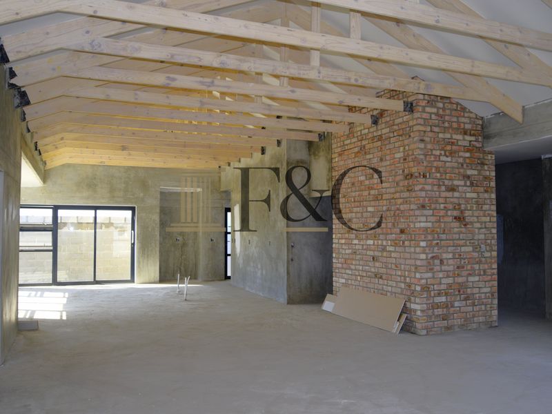 Spacious open-plan interior under construction, featuring exposed timber roof trusses, a prominent brick chimney breast, and large sliding glass doors.