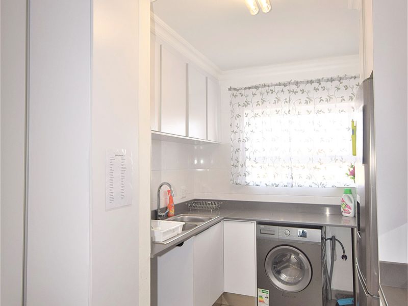 A functional laundry area featuring built-in white cabinetry, a stainless steel sink, tiled flooring, and space for a washing machine.