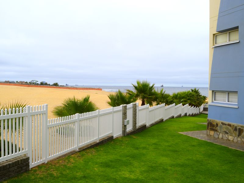 Rear exterior view of the apartment building featuring a well-maintained lawn, stone-clad facade, and a white picket fence overlooking the beach.