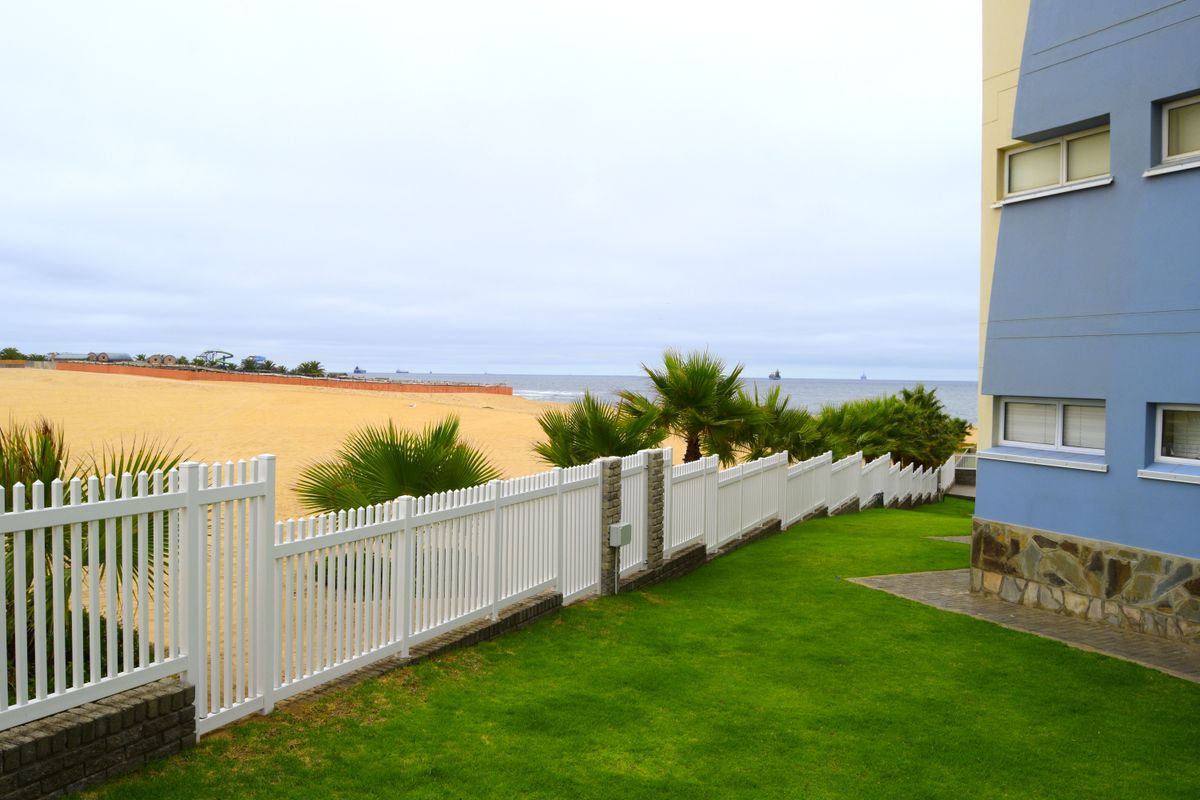 Rear exterior view of the apartment building featuring a well-maintained lawn, stone-clad facade, and a white picket fence overlooking the beach.