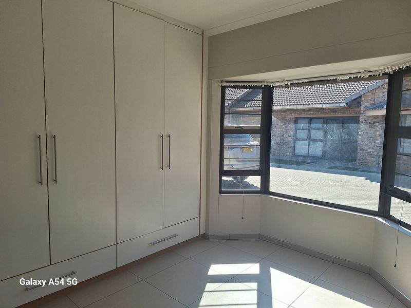 A bright bedroom featuring built-in floor-to-ceiling wardrobes, tiled flooring, and a large bay window overlooking the driveway.